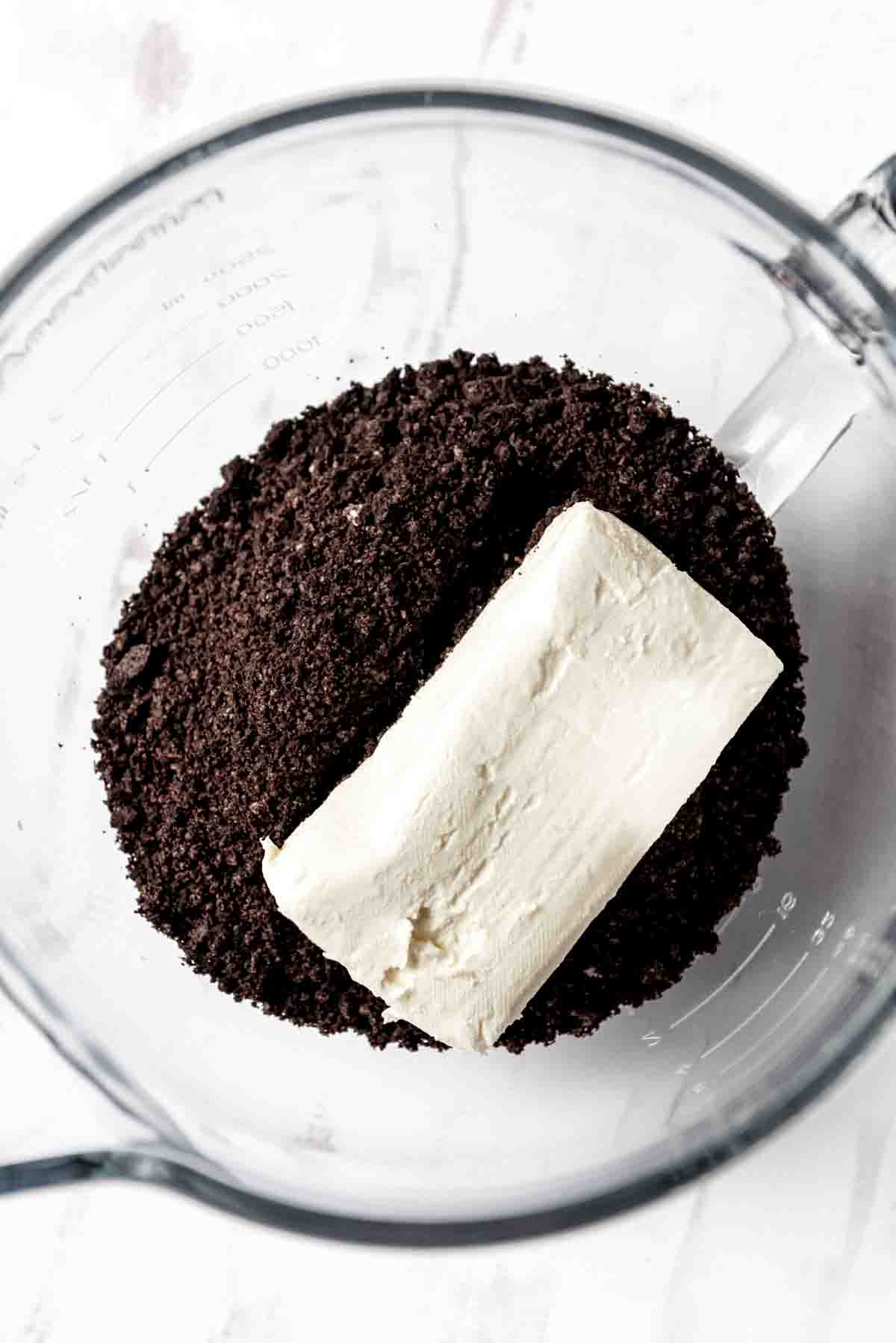 Adding softened cream cheese to Oreo cookie crumbs in a mixing bowl.