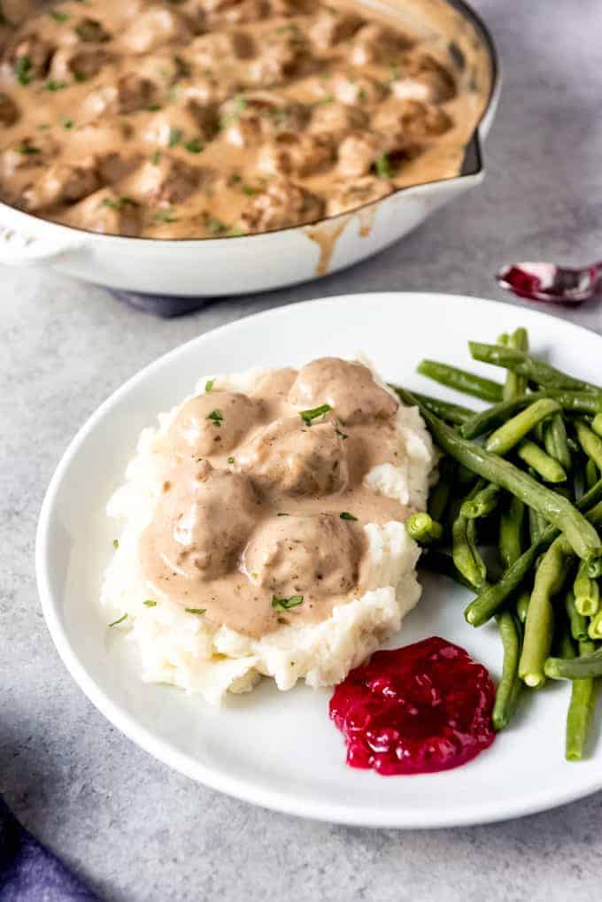 An image of a plate of Swedish meatballs served over mashed potatoes with the sauce, lingonberry jam, and green beans.