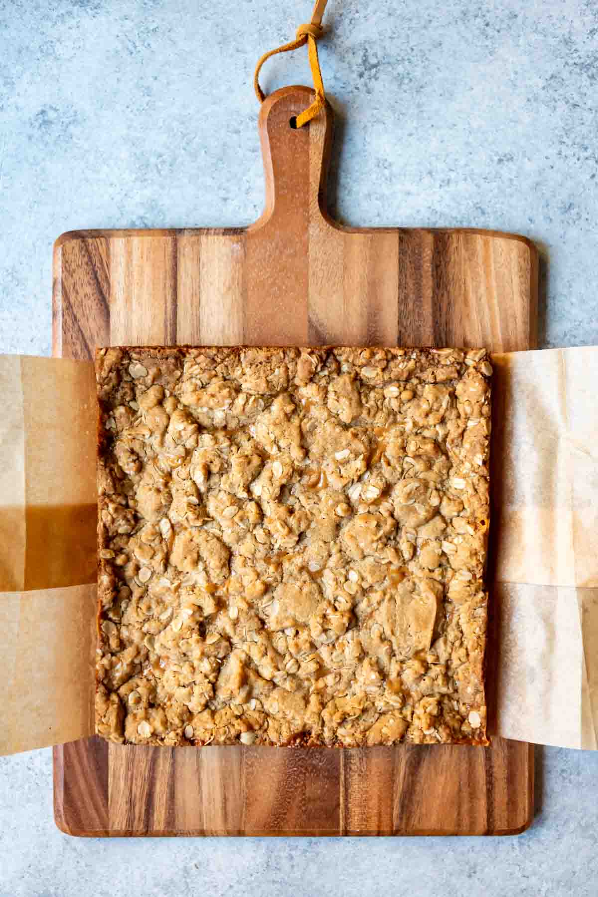 An image of carmelita bars in a parchment paper sling on a cutting board before being cut into squares.