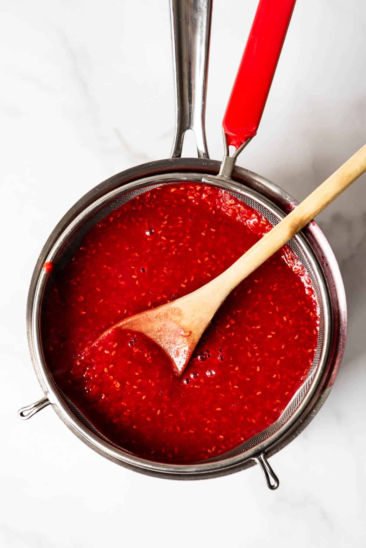 An image of pureed raspberries and sugar being pushed through a fine mesh strainer.