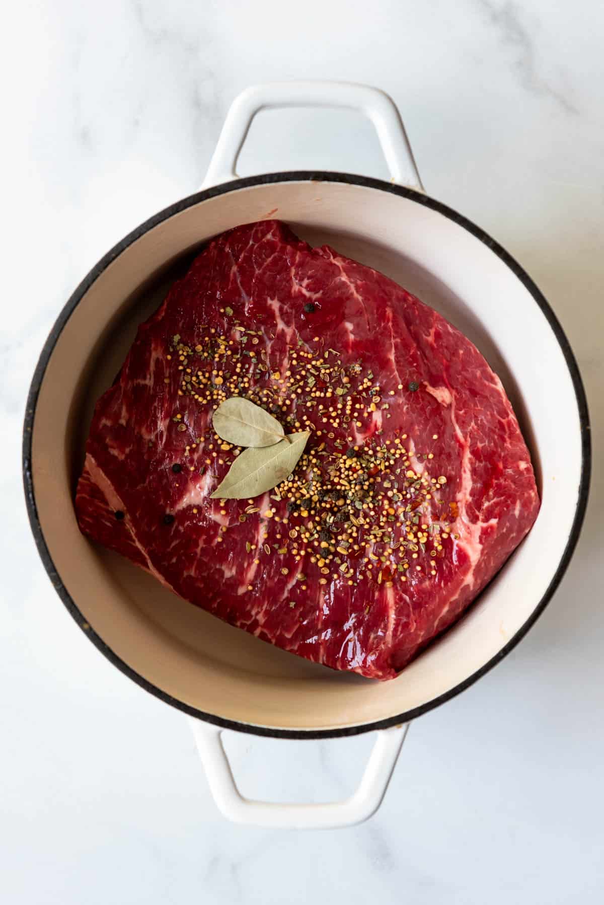 An image of a piece of marbled beef brisket in a dutch oven with pickling spices and bay leaves.