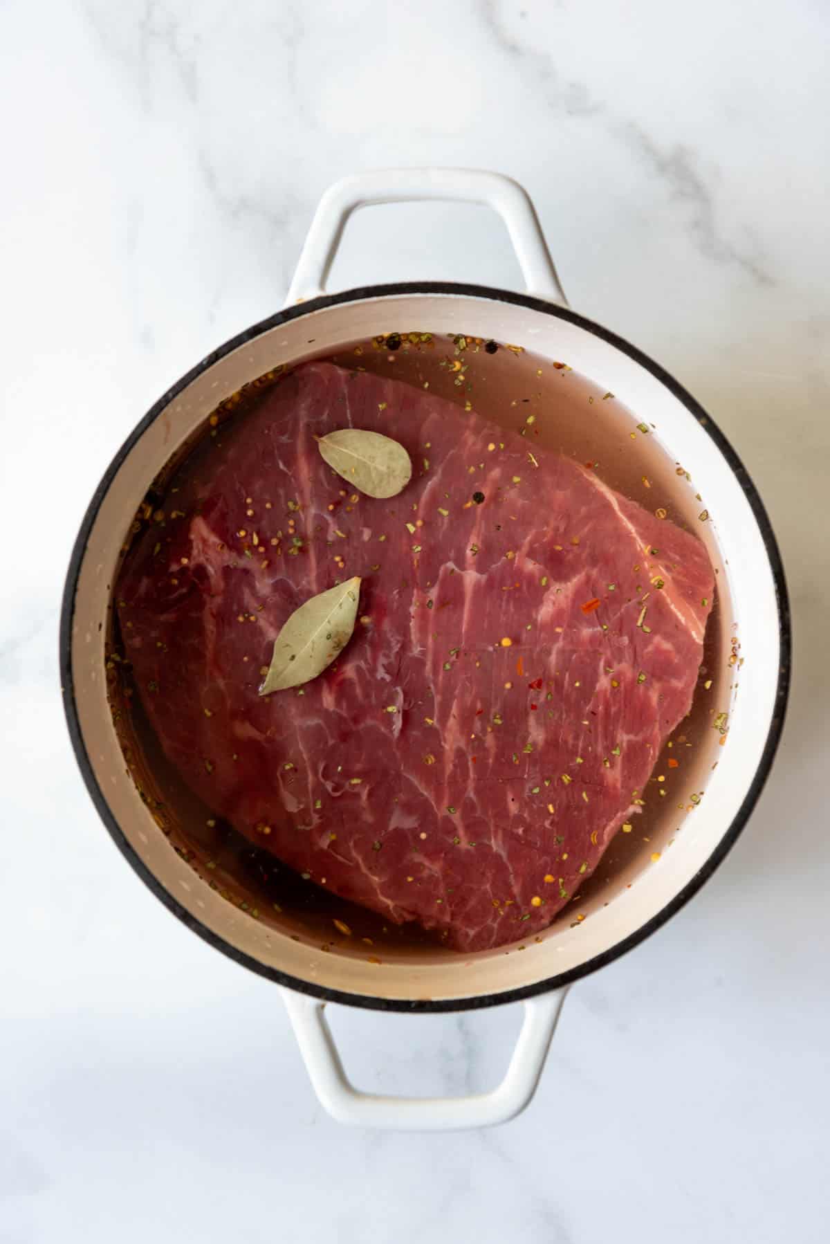 An image of a piece of marbled beef brisket in a dutch oven with pickling spices and bay leaves.