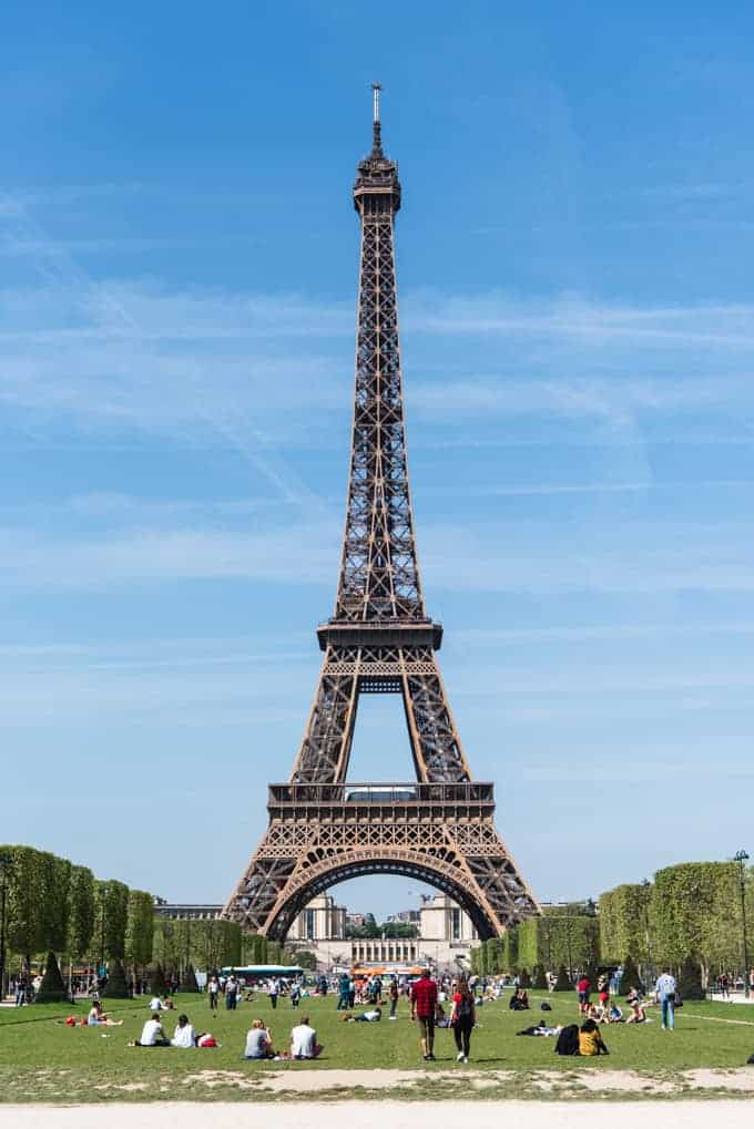 An image of the Eiffel Tower with people relaxing on the grass in front of it.