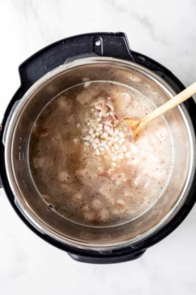 An image of an instant pot filled with small white beans, bacon, water, and spices.
