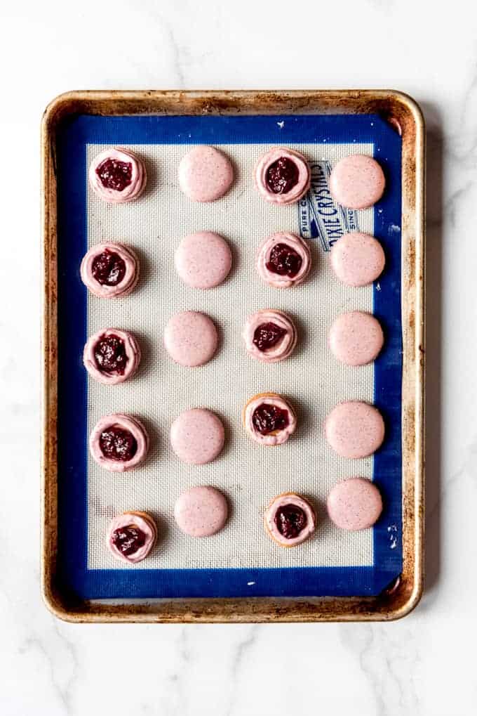 An image of macaron shells on a baking sheet being filled with buttercream frosting and jam.
