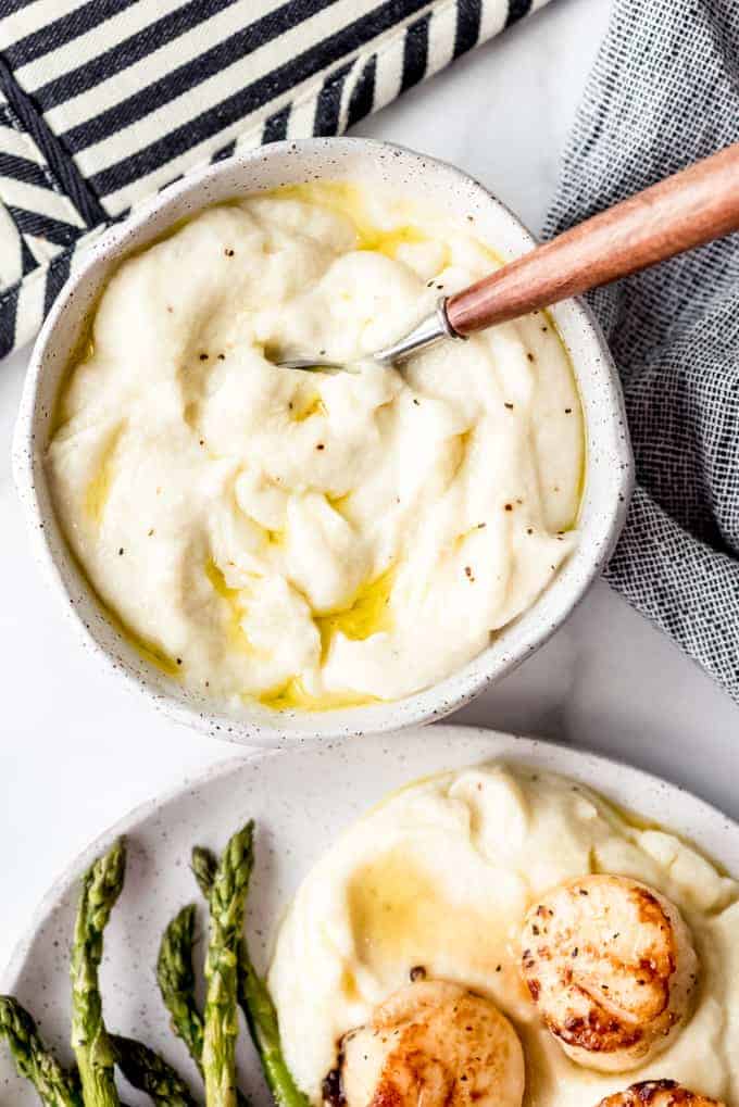a spoon  in a bowl of mashed cauliflower next to a plate with scallops and asparagus