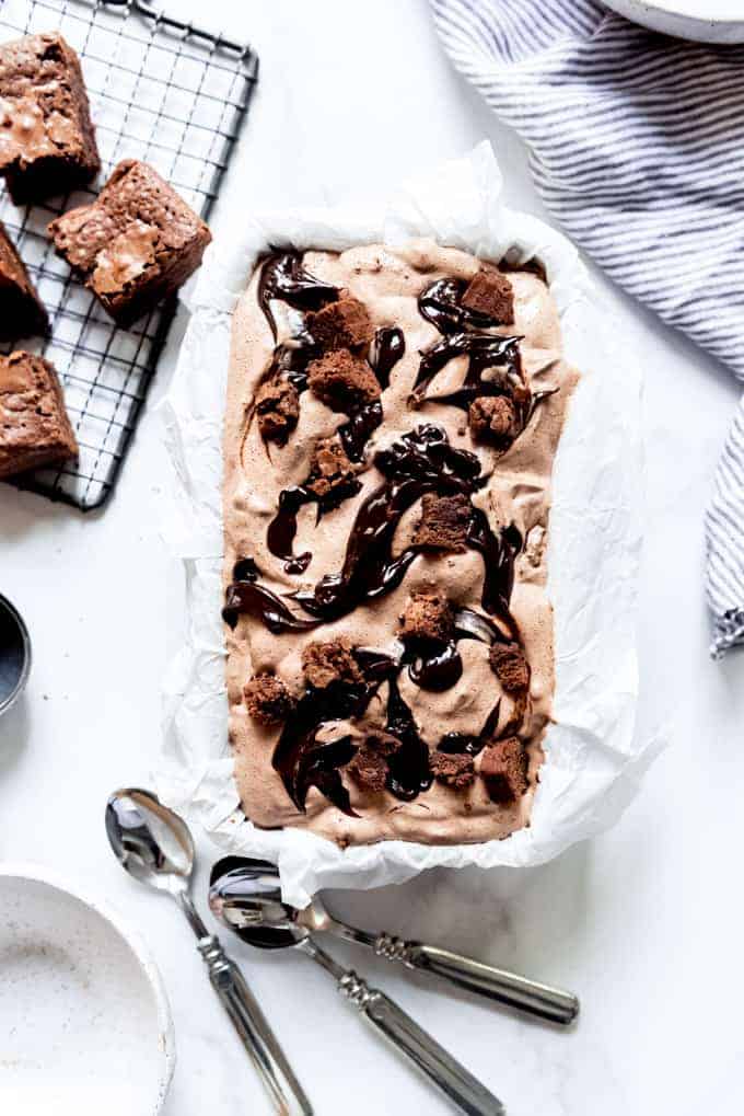 A bread pan filled with homemade chocolate ice cream with brownie pieces and a fudge swirl next to spoons and more brownies.