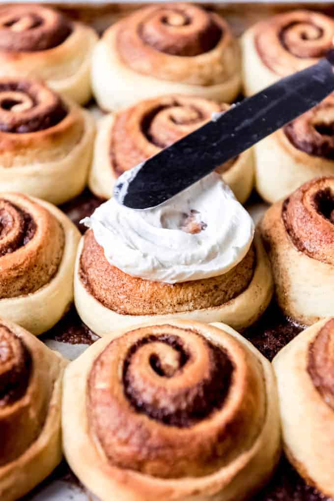 An image of frosting being spread on warm homemade cinnamon rolls.