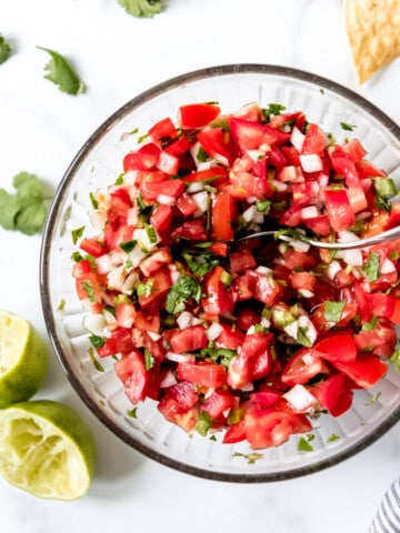 an aerial view of pico de gallo in a bowl with lime wedges and tortilla chips and cilantro around it