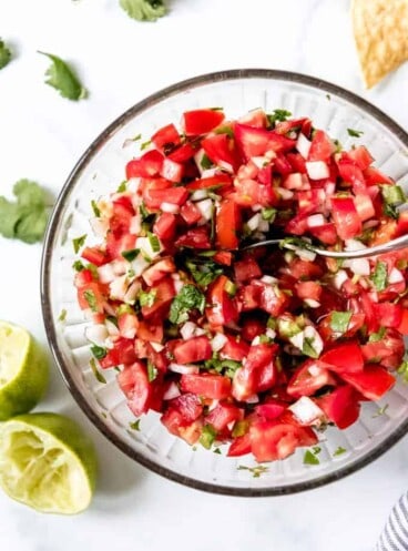 an aerial view of pico de gallo in a bowl with lime wedges and tortilla chips and cilantro around it