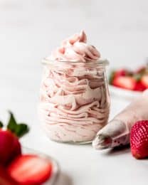 An image of strawberry frosting in a glass jar next to a piping bag with a decorative tip.