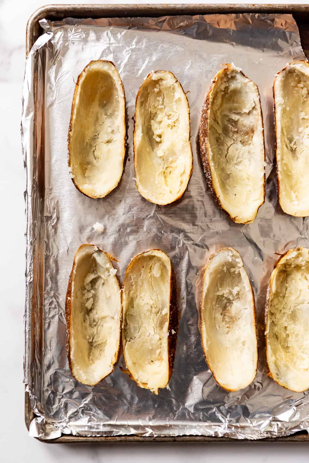 An image of hollowed out potato skins on a foil-lined baking sheet.