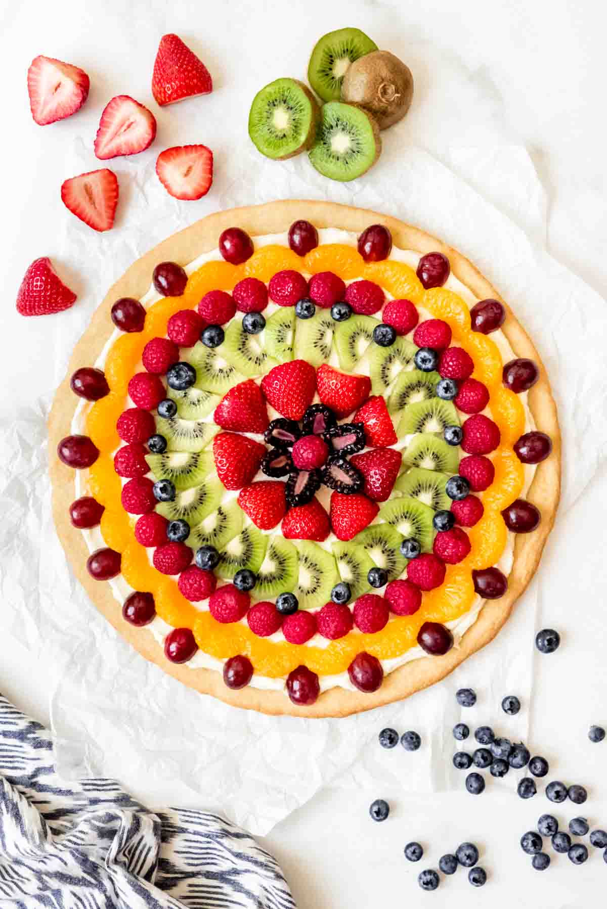 An image of a fruit pizza decorated with fresh kiwi, raspberries, strawberries, grapes, blueberries, and mandarin oranges.