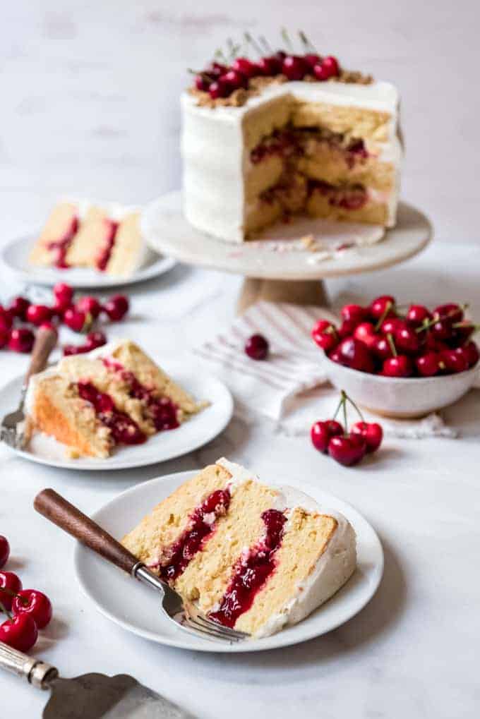 Slices of cherry crisp cake on white plates next to a bowl of cherries and the remaining cake on a cake stand.