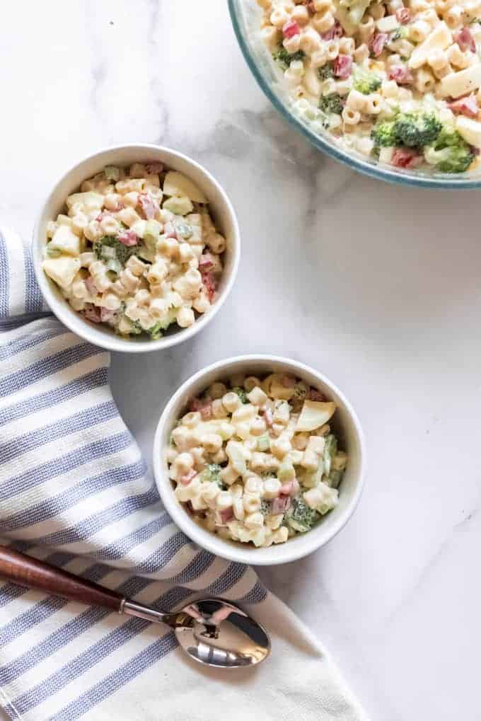 Two white bowls with servings of cold macaroni salad next to a larger serving bowl, a striped napkin, and spoon.