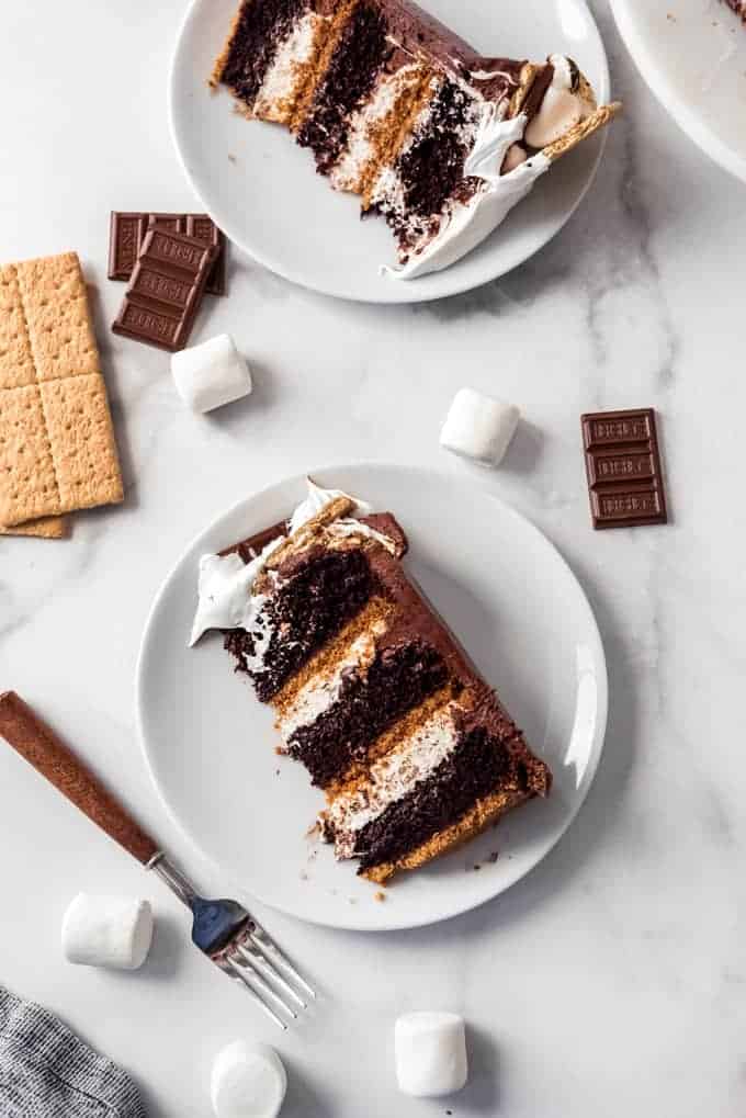 Slices of three-layer s'mores cake on white plates next to a fork, chocolate bars, graham crackers, and marshmallows.