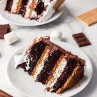 Two slices of smores cake on white plates with scattered chocolate, graham crackers, and marshmallows