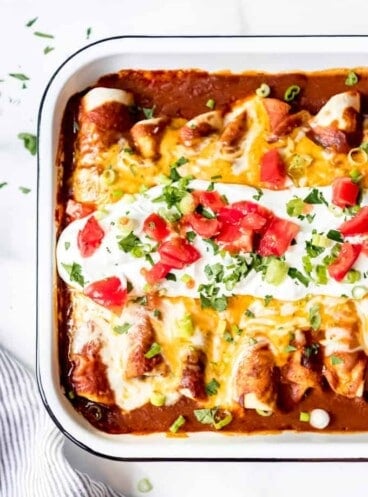 A white baking dish with beef enchiladas inside and garnished with sour cream and extra toppings