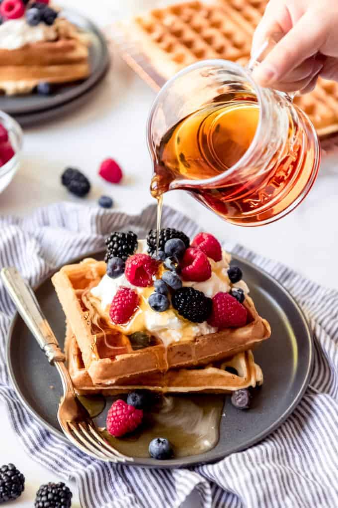 An image of maple syrup being poured over sourdough waffles topped with berries and whipped cream.
