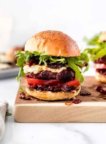 A burger topped with bacon jam, arugula, cheese, and sliced tomatoes on a cutting board.