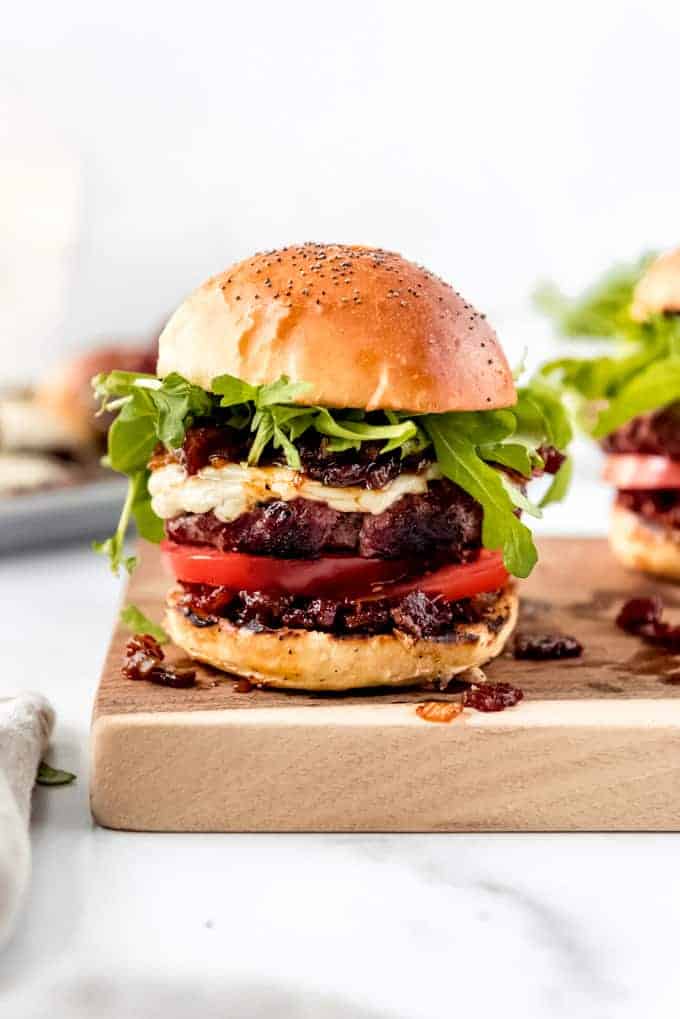 A burger topped with bacon jam, arugula, cheese, and sliced tomatoes on a cutting board.