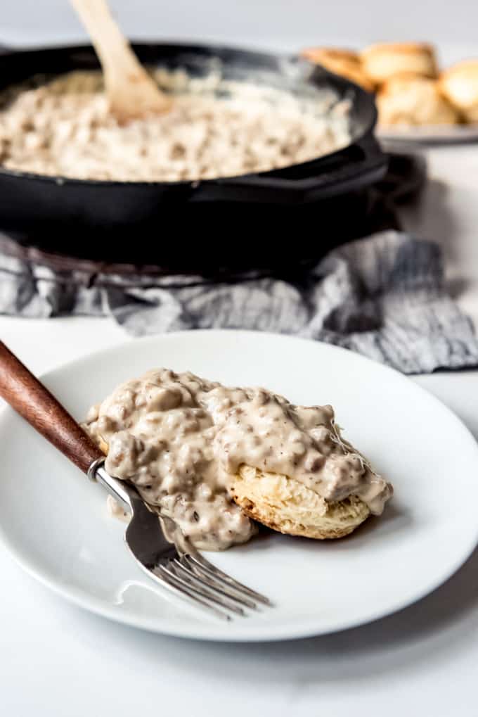 Buttermilk biscuits covered in sausage gravy on a white plate with a fork next to a cast iron skillet.