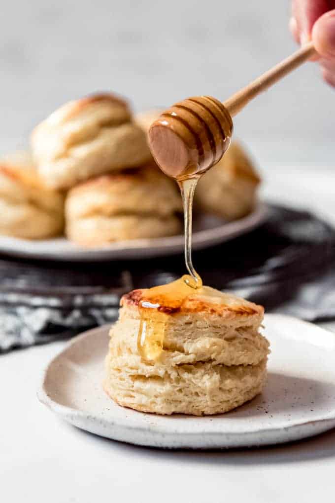 A honey dripper dripping honey over a buttermilk biscuit on a plate.