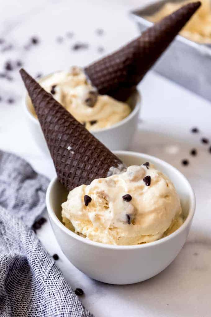 Two white bowls with scoops of chocolate chip cookie dough ice cream and chocolate sugar cones in them.