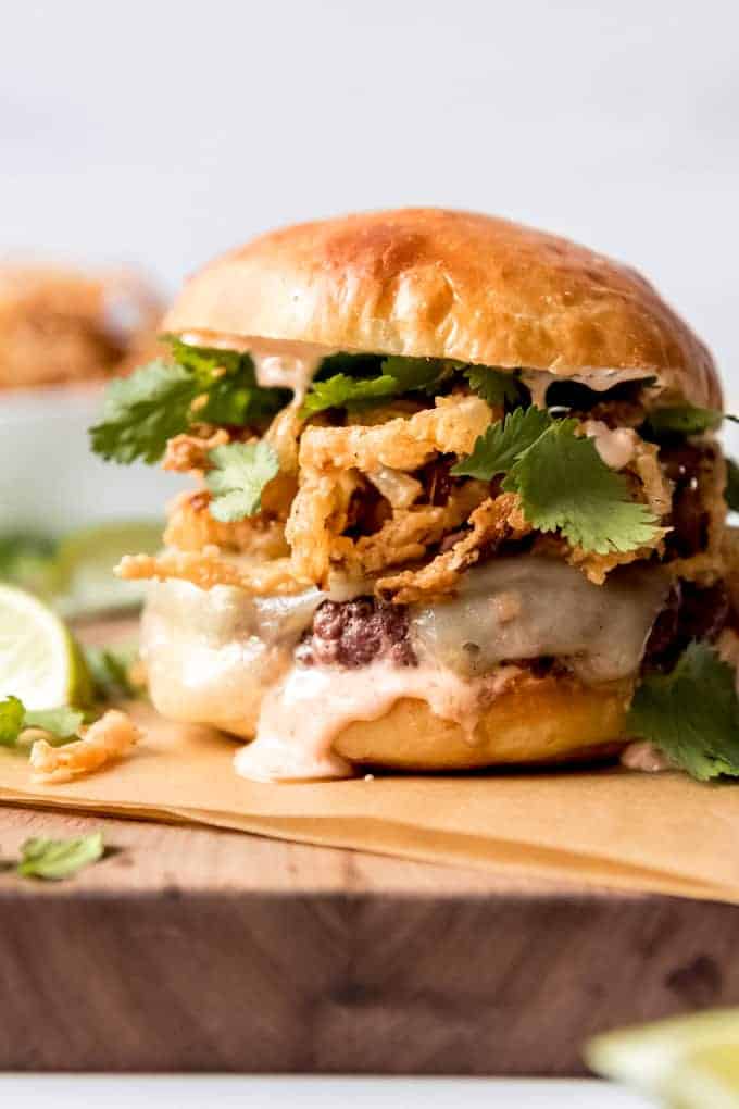 A burger topped with fried onion strings, sriracha aioli, and cilantro leaves on a wooden cutting board.