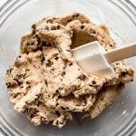 A glass bowl with a white spatula mixing mini chocolate chips into cookie dough.