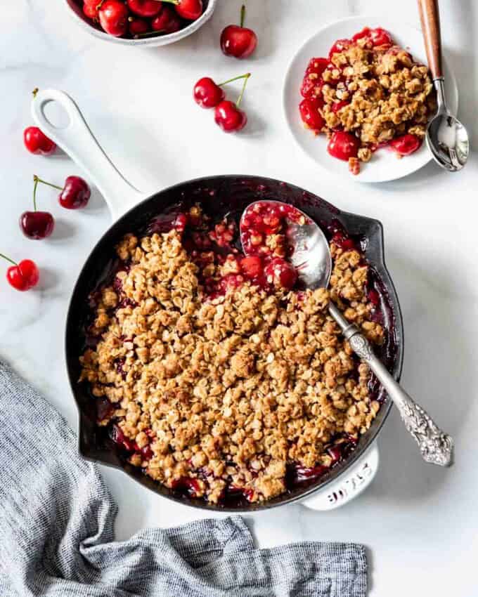 A large cast iron skillet with crisp oat topping over cherry pie filling next to some fresh cherries and a white plate.