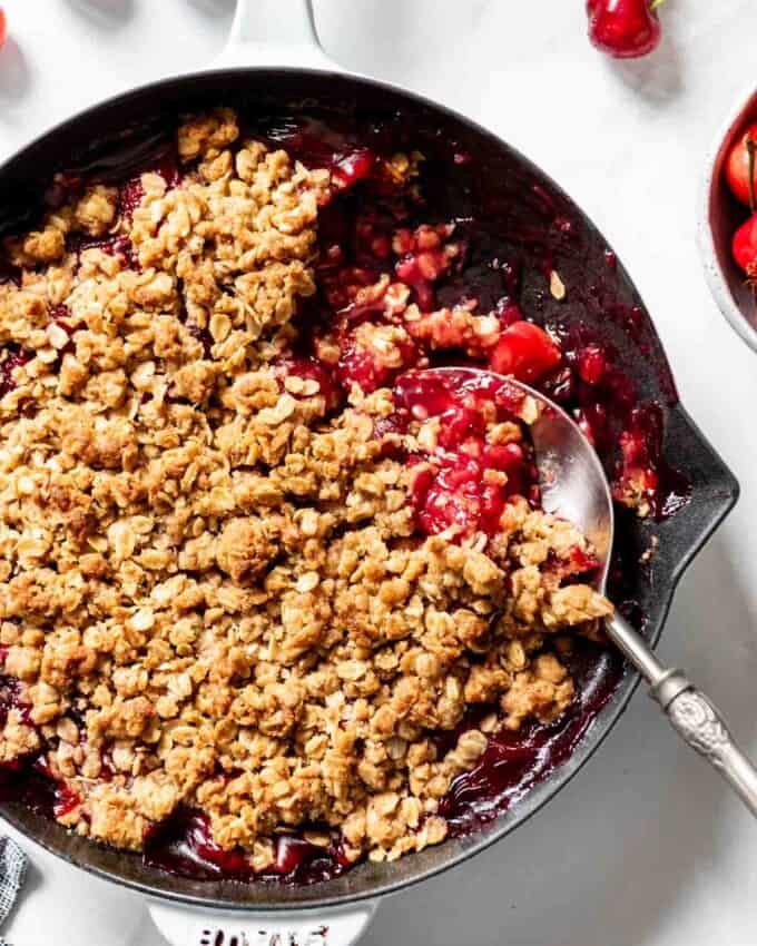 A serving spoon scooping out a portion of cherry crisp from a cast iron skillet.