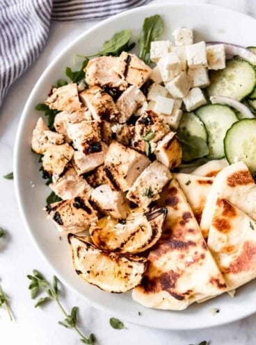 Grilled pieces of marinated chicken on a white plate with feta, pita, and cucumber slices.