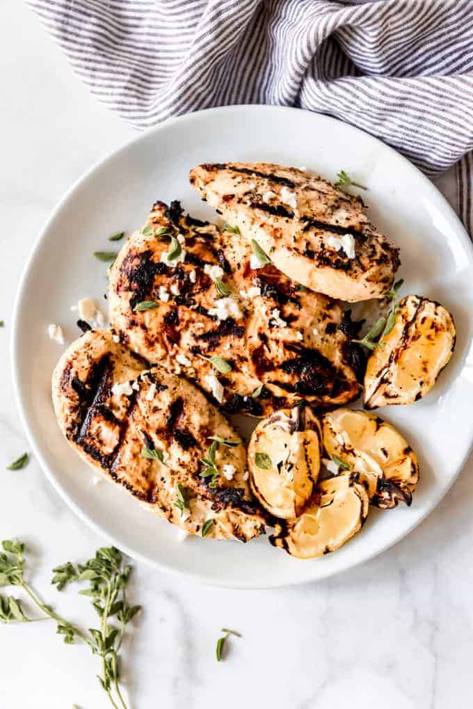 Grilled chicken breasts on a plate with lemon wedges, feta, and fresh oregano.
