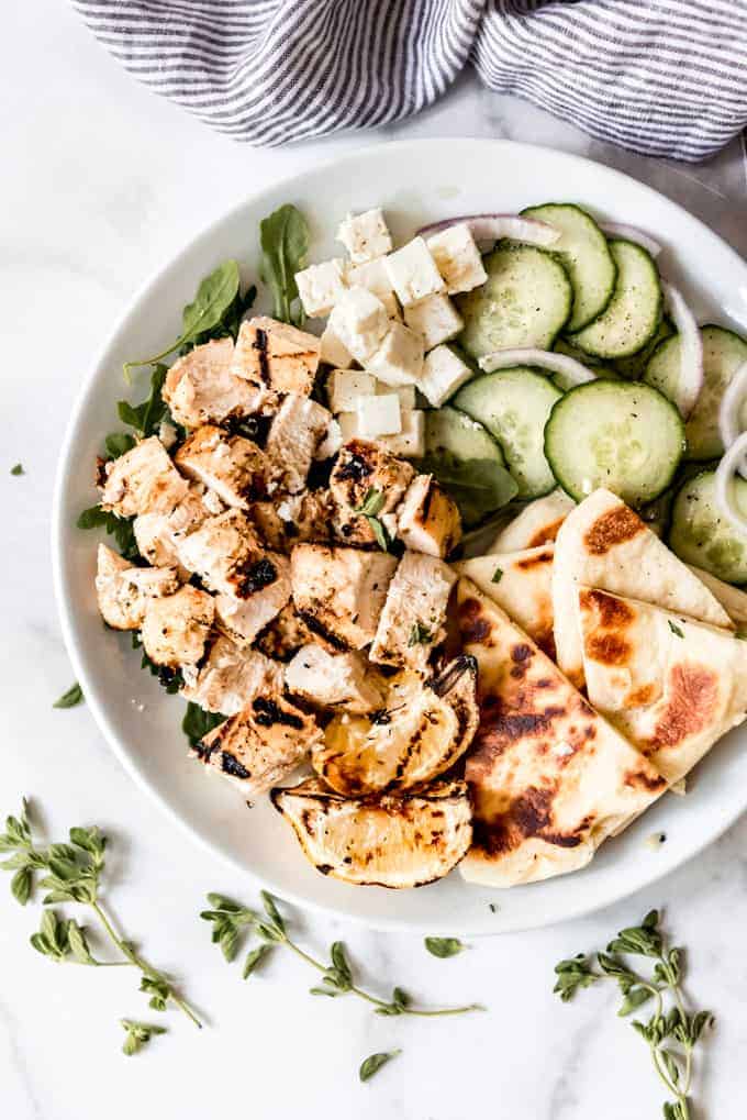 A white plate with grilled marinated chicken with sides of cucumber salad and pita bread.