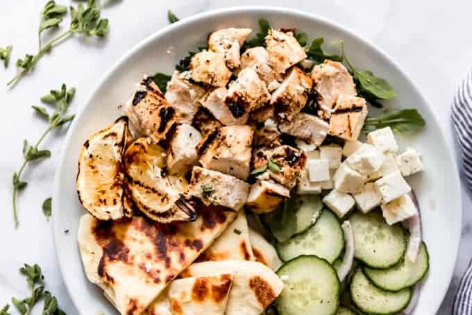 A white plate with chunks of grilled chicken, feta cheese, cumber slices, pita bread, grilled lemon wedges, and arugula.