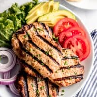 Grilled turkey burger patties on a white plate with sliced tomatoes, red onions, sliced avocado, and green leaf lettuce.