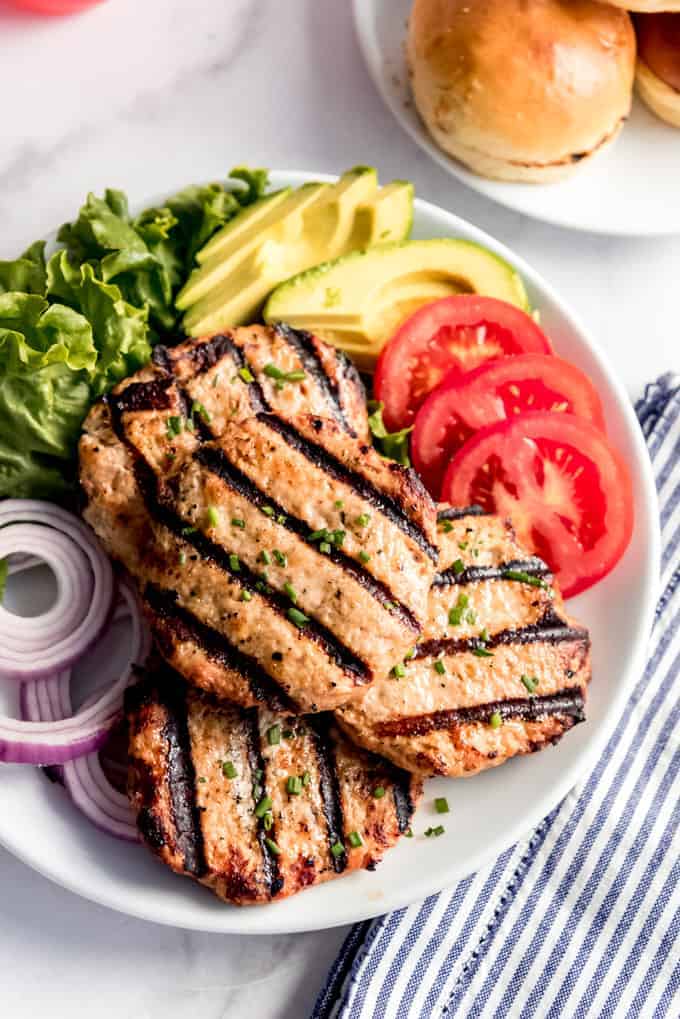 Grilled turkey burger patties on a white plate with sliced tomatoes, red onion, sliced avocado, and green leaf lettuce next to a striped cloth napkin.