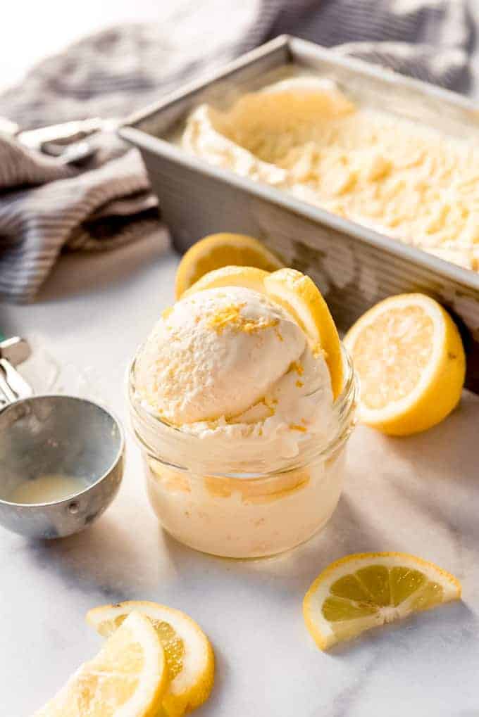 Lemon ice cream in a glass bowl next to sliced lemons and an ice cream scoop.