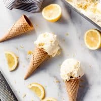 Scoops of lemon ice cream on sugar cookies on a marble surface surrounded by sliced lemons.