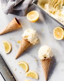 Scoops of lemon ice cream on sugar cookies on a marble surface surrounded by sliced lemons.