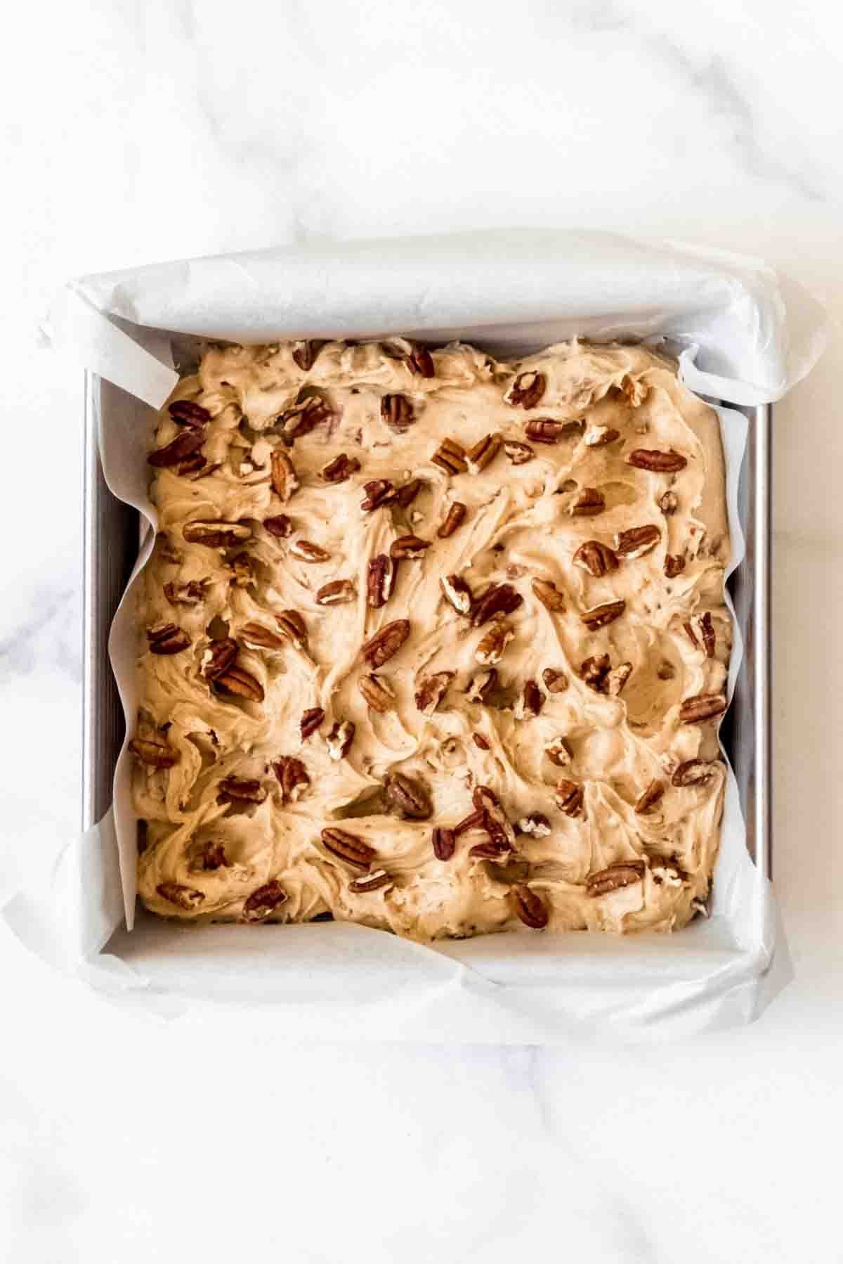 Maple pecan blondie batter in a square baking dish lined with parchment paper and chopped pecans sprinkled on top.