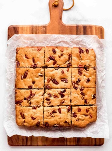 Blondies cut into 12 squares topped with chopped pecans on a cutting board.