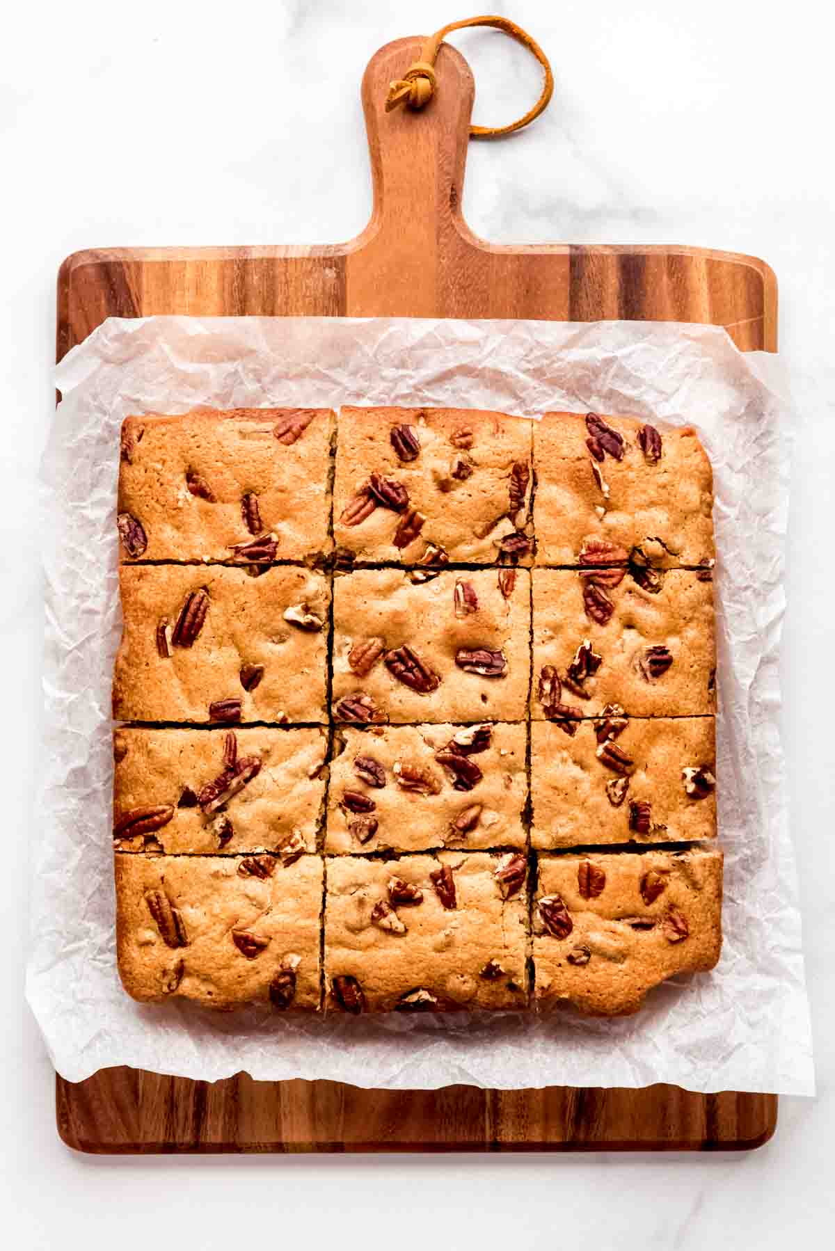 Blondies cut into 12 squares topped with chopped pecans on a cutting board.