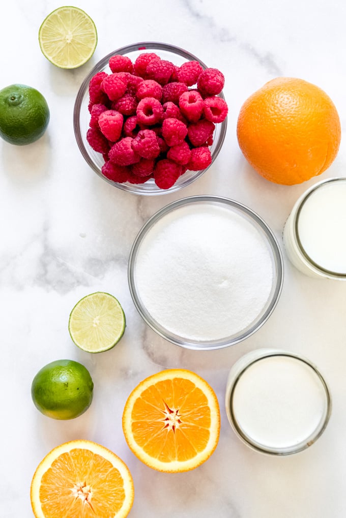 Bowls of raspberries, sugar, cream, and milk, next to oranges and limes.