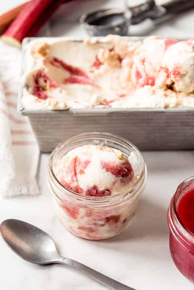 A glass dish with a scoop of rhubarb crumble ice cream in it in front of a container with the rest of the batch of ice cream.