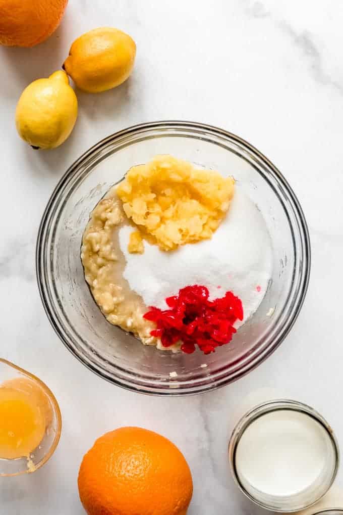 A bowl with mashed banana, crushed pineapple, sugar, and chopped maraschino cherries next to oranges and lemons.
