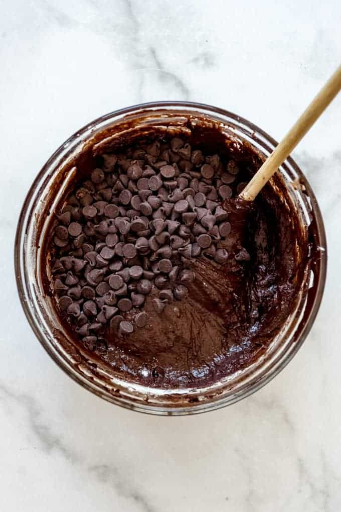 Brownie batter in a glass bowl with chocolate chips being stirred in with a wooden spoon.