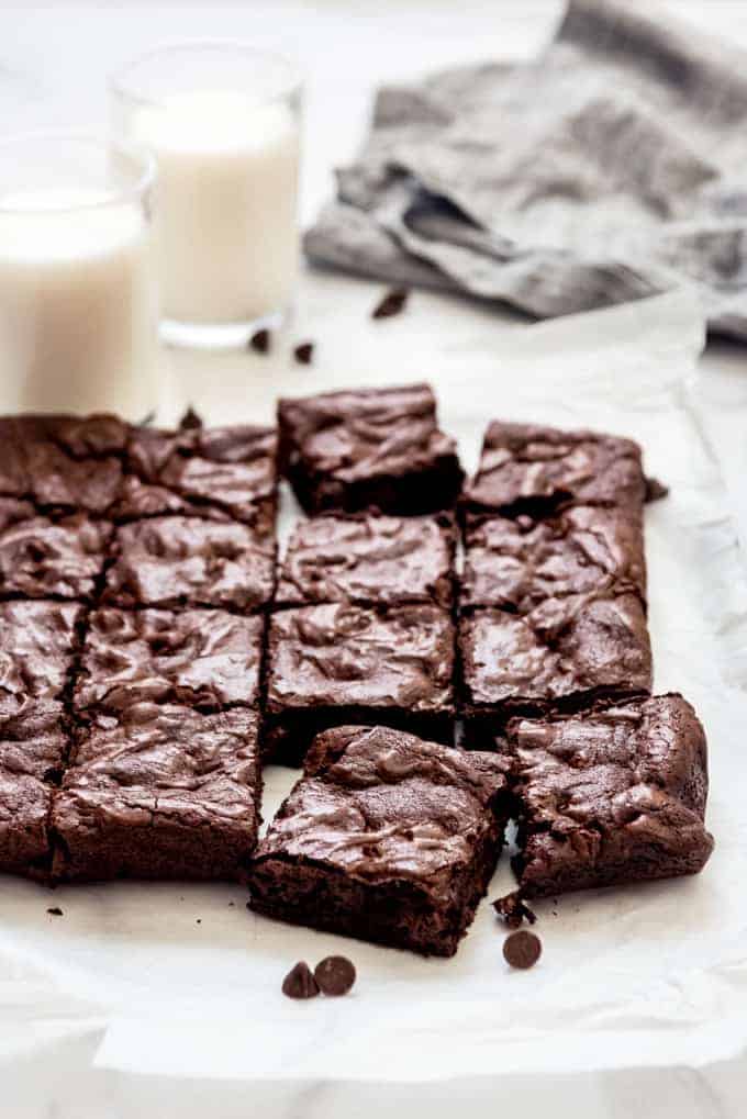 Brownie squares on parchment paper next to glasses of milk.