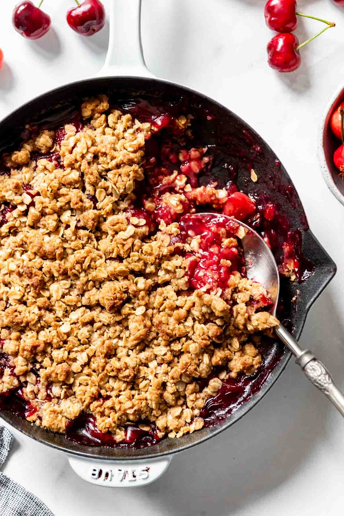 A serving spoon scooping out a portion of cherry crisp from a cast iron skillet.